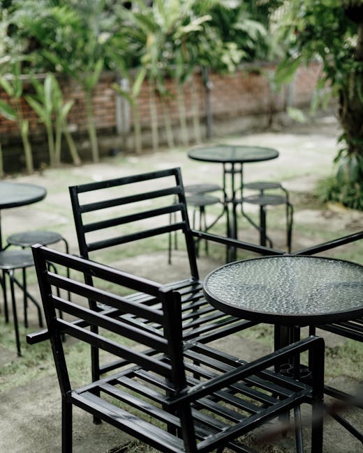 Serene outdoor café setting with empty black metal chairs and glass tables amidst lush green plants.