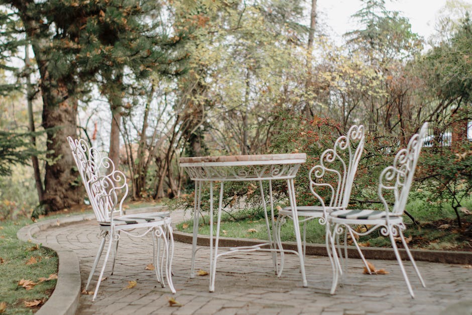 A tranquil outdoor patio setting featuring ornate white metal chairs and table surrounded by lush greenery.