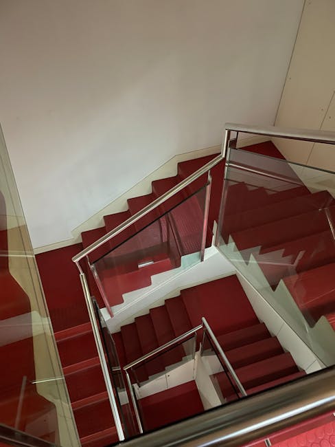 A high-angle view of a modern red-carpeted staircase, sleek metal handrails, and white walls.