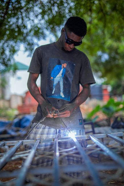 A man wearing a graphic t-shirt welds metal frames outdoors, shielded with sunglasses, showing sparks.