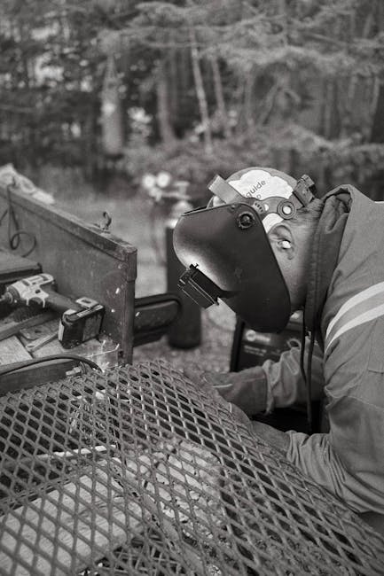 A welder wearing a protective helmet works on metal outdoors.
