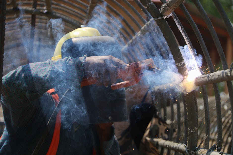 A welder wearing safety gear works with metal, producing smoke and sparks outdoors.