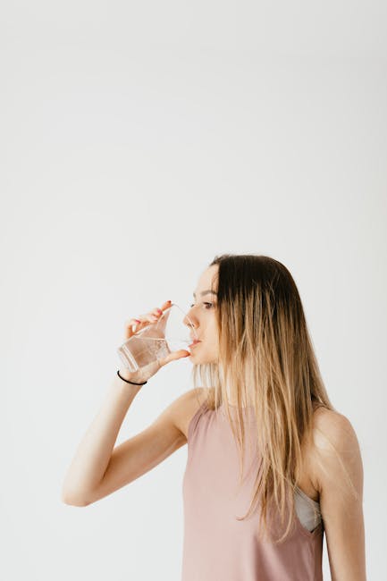Concentrated young fit female in sport top drinking water from glass while standing against white background