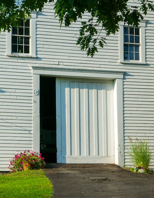 A vintage white barn with sliding doors, surrounded by lush greenery and flowers.